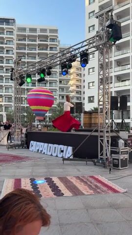 A male performer in traditional Sufi attire, including a tall, brown conical hat and a flowing red skirt, spins on a raised stage in front of large white letters spelling out "CAPPADOCIA." He is wearing a white shirt and dark shoes. The stage is lit by several green and blue stage lights mounted on a metal truss structure overhead. To the left of the stage, a large, colorful hot air balloon decoration is visible. In the background, a tall, modern white building with many balconies dominates the scene. The ground in front of the stage is paved with light-colored tiles, and a patterned rug is laid out on the ground. A few other people are present in the background, including one person standing near the hot air balloon. The performer is captured mid-spin, with his red skirt fanned out around him. The lighting suggests it is late afternoon or early evening.
