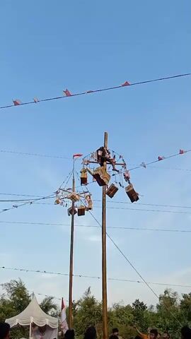 The video shows a low-angle view of a bright, clear sky with two tall wooden poles in the foreground, likely part of a traditional game or festival. Each pole has several tiers of wooden platforms or baskets attached, decorated with small red and white flags. On top of the poles, people are climbing and interacting with the baskets, some appearing to be retrieving items or engaging in a competition. Multiple strings with small red and white flags are strung between the poles and extend into the distance. Below the poles, a crowd of people is gathered, looking up at the activity. A white tent with red and white decorations is visible on the left side of the frame. The overall scene suggests a lively outdoor event, possibly in Southeast Asia, given the flags and the nature of the poles and activities.