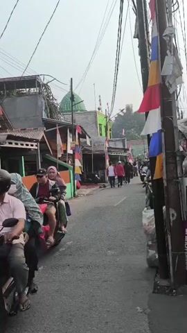 The video shows a street scene with several motorcycles passing by. On the left side of the street, there are buildings with colorful facades and flags. A green dome is visible in the background, suggesting a mosque. On the right side of the street, there is a utility pole adorned with flags. People are seen riding on motorcycles and walking along the street. The overall environment appears to be a residential or commercial area, possibly in Southeast Asia, given the presence of the mosque and the style of the buildings and vehicles. The weather seems to be overcast or hazy.