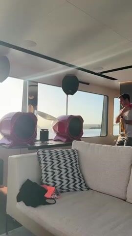 A man holds a bottle and appears to be pouring something from it into another container. He is standing near a large window with a bright sun visible outside. In the foreground, there is a couch with pillows and some clothing items. There are also large, red, drum-like speakers on a counter. Two black speakers are mounted on the ceiling. The scene appears to be indoors, possibly on a boat, with the water and sky visible through the window.