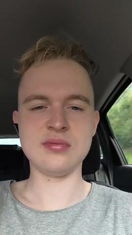 A close-up, front-facing shot shows a young white person with short, blonde hair styled upwards, and shaved sides. They are wearing a grey t-shirt and an earbud in their left ear. The person is in the driver's seat of a car, with the headrest and part of the car interior visible. Outside the car window, a blurry background of green trees and foliage suggests a natural, outdoor environment, possibly a road trip. The lighting is natural, indicating it is daytime. The person's expression is neutral, and they appear to be looking directly at the camera. The timestamp in the video is 00:00:00 to 00:04:04.