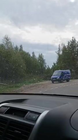 A blue van is seen driving on a paved road. The road is surrounded by trees and vegetation. The sky is cloudy. The van is driving away from the camera. The camera appears to be inside another vehicle.