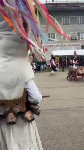 The video shows a festival scene in a town square, with people in traditional folk costumes and masks. The focus is on two individuals: one wearing a white furry costume with large bells attached around the waist and the other wearing a traditional Bulgarian outfit, including a red vest, white shirt, and a large headdress adorned with colorful ribbons. This person also has bells around the waist. In the background, other traditionally dressed figures are visible, with one group pushing a small wooden cart. People are gathered watching the spectacle, and a building with a sign in Bulgarian is in the background. The weather appears overcast.