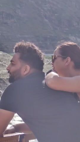 A man and a woman are in the foreground, with the man facing left and the woman facing right, resting her arm on his shoulder. They are both looking out at a river and a rocky, mountainous landscape in the background. The man is wearing a black t-shirt and has dark hair and a beard. The woman is wearing sunglasses and has her hair pulled back. The lighting suggests it is daytime and sunny.