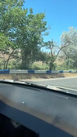 The video shows a view from inside a car. The dashboard is visible in the foreground, with a cigarette pack on the passenger seat. The car is moving along a road with a guardrail. In the background, there are mountains, trees, and a clear blue sky. The road appears to be in a rural or mountainous area. The overall impression is of a scenic drive on a sunny day.