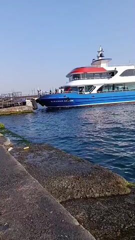 A bright blue and white passenger ferry is docked at a pier. The ferry has multiple levels, a red top deck, and large windows. The name "S. HASAN ALİM" is visible on the side of the ferry in white lettering. The ferry is positioned to the right of the frame, with its bow facing left. The water in front of the ferry is choppy and blue. In the foreground, a concrete pier with rough texture and some green moss is visible, running diagonally from the bottom left to the bottom right of the frame. The sky is clear and blue. No people are clearly visible on the deck of the ferry, though figures might be present near the railing. The overall scene depicts a ferry waiting at a dock on a clear day.