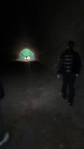 Two individuals are walking through a dark tunnel towards the light at the other end. The tunnel appears to be made of stone or brick. A vehicle with headlights on can be seen at the tunnel's exit. The person in the foreground is wearing a dark long-sleeved shirt, greenish pants, and a dark cross-body bag. The other person is wearing a dark puffer jacket and dark pants. The ground of the tunnel appears uneven and covered with debris.