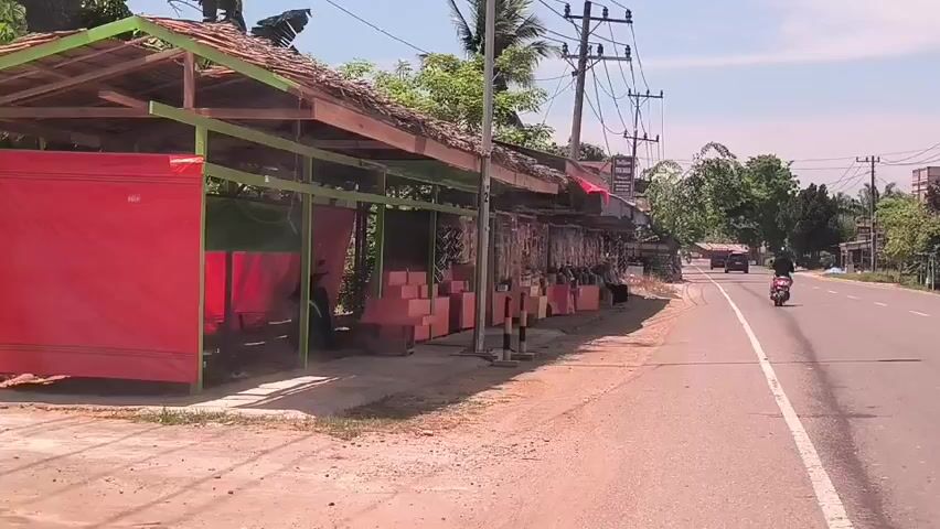 The video shows a straight road in a rural area on a sunny day. Along the side of the road is a row of simple structures, likely market stalls or shops. These structures have wooden frames, some with thatched roofs and supports painted green. The stalls display various items for sale, including what appear to be packaged goods, and some are draped with red fabric. A motorcycle travels on the road, and a car is visible further down the road. Power lines run alongside the road, supported by wooden poles. Trees and other vegetation are scattered throughout the scene, and the sky is blue with some scattered clouds.