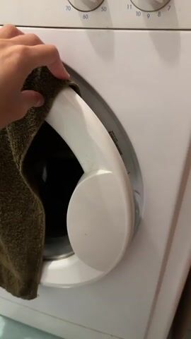 A person's hand is seen holding a green towel over the open door of a washing machine. The towel is partially obscuring the view inside the washing machine. As the video progresses, the towel is moved aside, revealing a brown cat lying inside the drum of the washing machine. The cat is looking towards the camera with its ears perked up. The washing machine is white and appears to be a front-loading model. The environment appears to be a laundry room, with a tiled floor visible in the background.