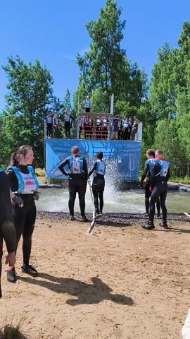 This media file captures an outdoor obstacle course event, likely a competition, taking place in Sertolovo, Russia. The scene is set on a sunny day, indicated by the clear blue sky and the distinct shadow of a person cast on the sandy ground. The participants, dressed in black wetsuits with distinctive bibs featuring numbers like "702" and "731", are engaged in a physical challenge.

In the foreground, a woman with blonde hair, wearing a similar wetsuit and bib, is lying down on the sand, looking towards the event. The main focus of the image is a group of participants in the water, pulling on a thick rope. Behind them, a blue structure with red netting serves as a part of the obstacle course, with more participants visible on top of it. The water appears to be a channel or a pool designed for the event. Spectators are gathered on a raised platform behind the blue structure, observing the competition. The surrounding environment is lush and green, with many trees suggesting a natural, possibly park-like, setting. A man in camouflage uniform is visible standing near the water's edge in the background. The overall atmosphere seems energetic and competitive. Visible text includes numbers on the participants' bibs and some text on the blue structure, which is partially obscured but appears to be in Russian. The time of day is clearly daytime, given the sunlight and shadows.