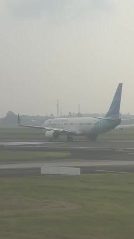 A white airplane with a distinctive blue and green tail, bearing the "Garuda Indonesia" logo on its fuselage, is featured in the video. The aircraft is situated on an airport taxiway or runway, viewed from a slightly elevated perspective. In the foreground, a red and white striped windsock is visible, indicating the presence and direction of wind. The entire scene is bathed in a soft, hazy light, suggesting either early morning or late afternoon with some atmospheric fog or mist. The ground consists of paved surfaces for aircraft movement, bordered by green grass. Distant structures and trees are partially obscured by the haze. The airplane appears to be stationary or moving very slowly, possibly waiting for clearance or taxiing to a gate/runway. The setting is an airport, with the provided context suggesting it is located in Pajang, Indonesia.