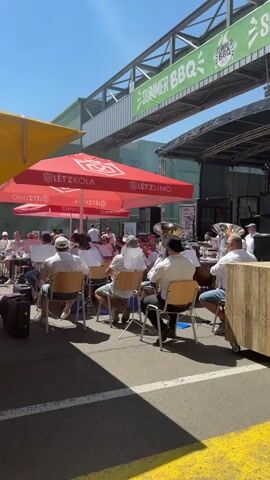 A brass band is playing music outdoors. The band members are seated in chairs, most of them wearing white shirts. Some are playing brass instruments like tubas and trumpets. They are positioned under red umbrellas with logos. In the background, there is a stage and a banner that says "SUMMER BBQ". The scene appears to be during the daytime, with bright sunlight.