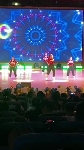 A group of five people, appearing to be young women, are dancing on a stage in front of a large, circular, blue and white illuminated screen. They are all wearing similar red tops with dark pants and are in coordinated poses. The stage floor is lit with white and green lights. Below the stage, in the foreground, an audience is seated in dark theater seats, with some audience members holding up cell phones. The audience is partially illuminated by stage lights and is facing the performers. There are balloons in front of the stage. The video is shaky and the lighting is dim in the audience area.