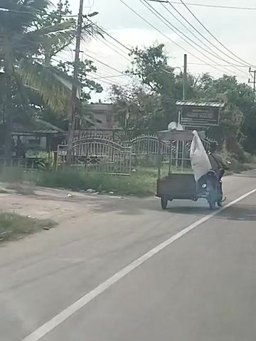 A scooter is parked on the side of a road. The scooter is dark colored and has a basket on the back. There is a light-colored tarp covering something in the basket. A sign is visible behind the scooter. The sign is dark and has white lettering. Trees and foliage are visible behind the sign. The road is paved and appears wet. The sky is overcast.