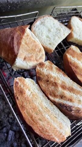 The video shows slices of bread being toasted over a fire. The bread is on a metal grill with a charcoal fire beneath it. Some slices are golden brown, while others are still white. The background is blurry but appears to be outdoors.