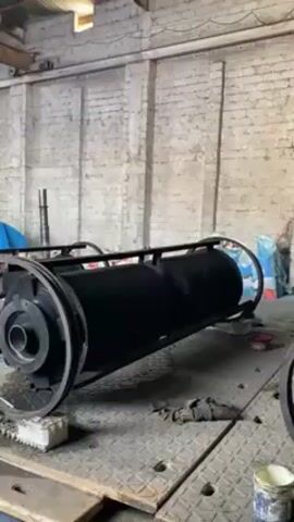 The video shows the interior of a large warehouse with bare brick walls. A large black cylinder-shaped object with wheels is positioned on the floor. A man wearing a white hat and black clothing is standing near the object. There is other equipment in the background, including a large blue object covered in plastic. The cylinder-shaped object appears to be newly painted.