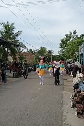 A parade is taking place on a paved road. A marching band, composed of young people in matching uniforms of turquoise tops and yellow skirts with white pants, is moving down the road. They are holding musical instruments and flags. In front of the marching band, a person in a military uniform is walking. On the sides of the road, spectators are watching the parade. Some spectators are standing, while others are sitting on motorcycles or in front of houses. There are trees and buildings visible in the background. The sky is cloudy.