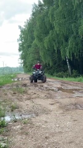 The video shows a young person riding a white and black ATV on a muddy, dirt road. The person is wearing a maroon jacket. The road is flanked by green trees and grass. The sky is overcast. The ATV is moving slowly over the rough terrain.