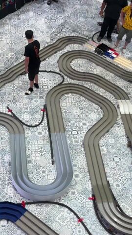 From a high-angle view, the video captures a slot car race on a complex, multi-lane track laid out on a floor with a patterned tile design. The track is primarily gray with segments of white, marked with red and blue accents. Several people are positioned around the track, observing the race. One person is wearing a black shirt and black shorts. Another has on a yellow patterned shirt. The slot cars move quickly along the track. There is an overall atmosphere of casual observation and interest in the race.