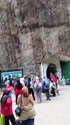 A group of tourists, predominantly women wearing hijabs and casual clothing, are gathered outside a large cave entrance carved into a rocky cliff face. Some individuals are wearing hats. A informational display board with images is visible to the left of the cave entrance. A green gate is also present to the right of the cave. The ground is paved with interlocking bricks. The tourists appear to be queuing or preparing to enter the cave. The overall atmosphere suggests a tourist attraction or historical site.