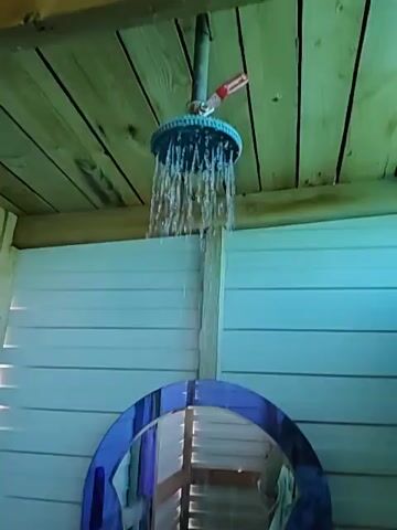 The video begins with a close-up view of a showerhead dispensing water. The showerhead is attached to a pipe that extends from a wooden ceiling. The shower stall appears to be outdoors, with white paneled walls and a wooden floor. A blue arched mirror is mounted on one of the walls, reflecting the interior of the shower stall. The reflection shows a person holding a camera or phone, presumably taking the video. The camera then pans down to show a person's foot wearing a white sandal with a colorful design. Water is seen falling onto the wooden floor. The scene is lit with natural light, suggesting it is daytime. The overall impression is that of an outdoor shower setup.