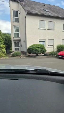 A dashcam view from inside a car shows a red Chevrolet Corvette C8 Stingray slowly driving past a light-colored, two-story house with several windows, some covered with horizontal blinds, on a sunny day. The Corvette has black rims and is driving on a paved road. The house is surrounded by green bushes and plants.