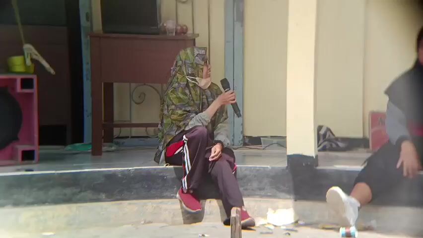 A person wearing a patterned hijab and glasses is sitting on steps holding a microphone and speaking. They are wearing a camouflage jacket over a gray shirt and dark pants with red and white stripes. Red shoes are on their feet. To the right, another person in a black hijab is sitting on the steps. In the background, there is a table with a laptop and some items on it. To the left, a pink speaker is visible. The ground is paved and appears to be outdoors.