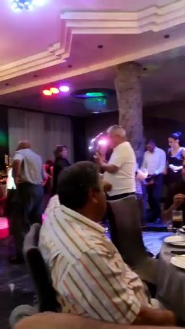 The video shows a dimly lit indoor event space, possibly a banquet hall or restaurant, with multiple people present. The lighting is colorful and dynamic, with blue, green, and yellow lights projected onto the ceiling and walls. In the foreground, a man wearing a striped shirt is seated at a table, facing away from the camera. In the midground, several people are interacting. A man and a woman are dancing together. Another man in a white shirt is standing nearby. Further back, a group of people are gathered, some of whom appear to be dancing. A woman in a black dress is visible, and another person is singing. In the background, a man is standing near a doorway, holding a phone. Tables are set with drinks and food. The overall atmosphere suggests a celebration or party.