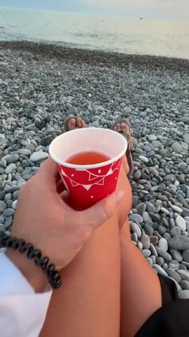 A first-person perspective video shows a hand holding a red cup filled with an orange beverage. The person is sitting on a pebble beach with their legs extended, wearing sandals. In the background, the ocean and a sunset are visible. The water is a muted orange, reflecting the colors of the sky. The sky transitions from soft pink and orange hues near the horizon to a deeper red where the sun is setting. There are a few birds flying in the distance above the water. The waves are small and gentle.
