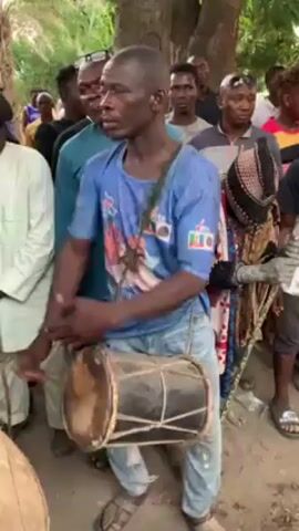A man is the primary focus of the video, vigorously playing a drum, suspended from his shoulder with a strap. He is surrounded by a crowd of onlookers in what seems to be an outdoor gathering. He is wearing a blue t-shirt and light blue jeans. His facial expression is intense, focusing on his playing. The other individuals in the video appear to be observing, some smiling and others with neutral expressions. One person to the right of the drummer is wearing what appears to be a tribal or ceremonial mask and outfit. The environment appears to be an open space with trees, suggesting a village or rural setting. The lighting suggests it is daytime. The ground is covered in dirt and loose gravel.