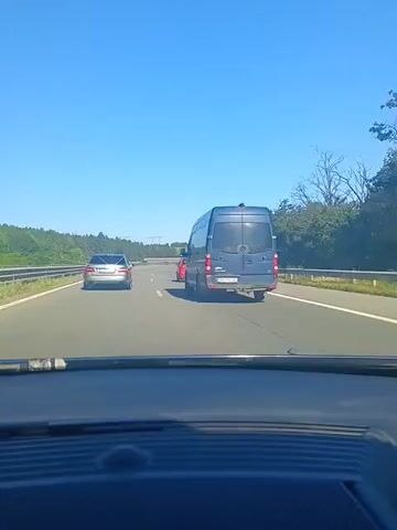 A dark blue Mercedes-Benz Sprinter van is seen from the perspective of a driver on a multi-lane highway. The van is in the right lane. In the left lane, a silver Mercedes-Benz sedan is ahead of the van, and a red hatchback is to the left of the silver sedan. The sky is clear and blue, and the road is bordered by green trees and vegetation. The van's license plate is visible but unreadable. The video is likely a dashcam recording.