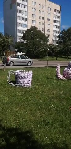 The video shows a sunny outdoor scene with a patch of green grass in the foreground. Two large, decorative objects made of white wire frames filled with what appear to be small, light pink or white stuffed items are placed on the grass. To the left is a large mug-shaped object with a handle. To the right is a teapot-shaped object with a spout and handle. In the background, there is a road with a silver car driving on it and several multi-story buildings in the distance. Trees are visible between the road and the buildings. The camera pans slightly to the right and zooms out slightly.