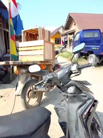 The video pans across a rural outdoor scene on a sunny day. In the foreground, the handlebars and front of a black motorcycle are visible. To the left, a motorcycle with a wooden crate attached to its rear rack is parked. Behind the motorcycles, a blue pickup truck is parked, followed by other vehicles including a white van and another blue pickup truck. In the background, wooden houses with corrugated metal roofs are visible, along with colorful flags. A person walks across the frame towards the right. The ground is dirt and gravel.