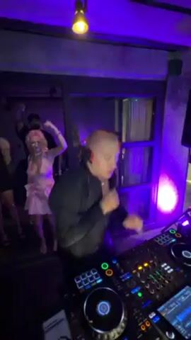 A DJ in a black hoodie mixes music at a party. The DJ is a light-skinned male with a shaved head. He is wearing headphones and looking at his equipment. In the background, people are dancing to the music. The people in the crowd are a mix of men and women, most wearing dark clothing. The party appears to be outdoors at night with lighting.  Some are holding up their phones.