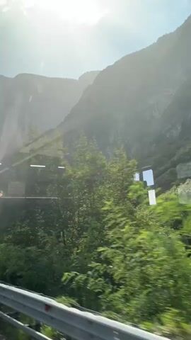 The video shows a view from a vehicle moving alongside a road, with a guardrail visible in the foreground. Beyond the guardrail, lush green trees and bushes line the side of the road. In the midground, a dense forest covers the slopes of a large, rugged mountain. The mountain is partially obscured by a bright sun and hazy light, creating a lens flare effect across the video. The overall impression is a scenic drive through a mountainous, forested area on a sunny day.