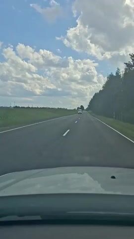The video captures a point-of-view shot from inside a vehicle traveling on a highway. The road ahead is asphalt with white lane markings. On the left side of the road, there is a vast green field. On the right side, a dense line of trees runs along the edge of the road. The sky above is blue with scattered white, fluffy clouds. A white vehicle, possibly a van or RV, is visible in the distance, traveling in the same direction. The dashboard of the vehicle from which the video is taken is partially visible in the foreground.
