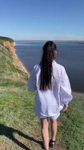 A woman with long dark hair stands on a grassy cliff overlooking a wide body of water. She wears a white oversized shirt, white shorts, and black sandals. The sky is blue and clear. The land on the other side of the water appears distant. The woman turns, smiles at the camera, then turns away again, and walks a short distance along the cliff's edge.