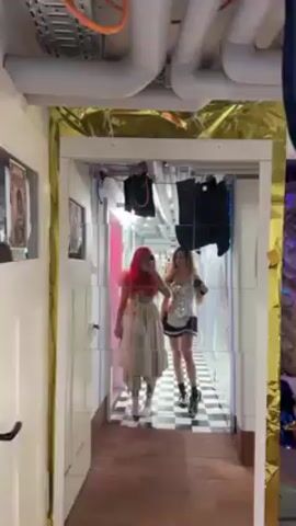 Two young women stand side by side in a hallway, visible via a segmented mirror. The woman on the left wears a long, frilly, off-white dress, white sneakers, and a bright red wig. She holds a phone in her left hand, presumably taking the photo. The woman on the right is dressed in a maid outfit with a short black skirt, a silver corset top, and black boots. The hallway has a black and white checkered floor, with white walls decorated with silver metallic textures. Overhead, exposed pipes and hanging objects are visible. A pink light illuminates a door on the left side of the hallway.