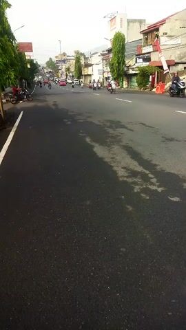 The video begins with a view of an asphalt road from a low angle, showing the texture of the freshly paved surface with some damp patches. The road is lined with trees on the left and buildings on the right. A white line marks the edge of the road. In the distance, the road curves and appears to be a busy street with several motorbikes and some cars. There are people on the motorbikes, and some are parked on the side of the road. The buildings on the right are a mix of commercial establishments, with some appearing to be shops or houses. There's a red and white flag pole visible near one of the buildings. The sky is overcast, suggesting it might be early morning or late afternoon. The overall scene depicts a typical urban street environment with ongoing activity.
