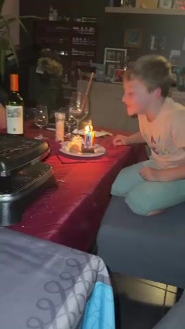 A young boy sits at a table with a red tablecloth. There's a small dessert plate in front of him with three lit birthday candles. He blows out the candles. There are wine glasses, a salt shaker, and other items on the table. A raclette grill is partially visible on the left. The boy is wearing a peach-colored shirt with "OLA AMIGOS" printed on it, and light green pants. He is sitting on a gray cushion on what appears to be a sofa. In the background, there's a shelf with various bottles and decorations. The room is dimly lit.
