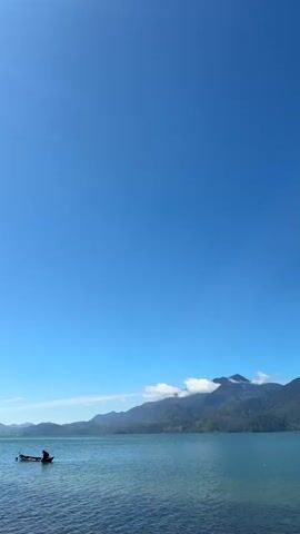 The video captures a serene landscape with a bright blue sky dominating the upper portion of the frame. A calm, blue body of water stretches across the lower half, with gentle ripples on its surface. In the distance, a range of lush, green mountains rises, their peaks partially obscured by a few wispy white clouds. To the left of the frame, a small, dark boat is visible on the water. A solitary figure is seated in the boat. The overall atmosphere is peaceful and tranquil, suggesting a clear, sunny day in a natural, mountainous coastal environment.