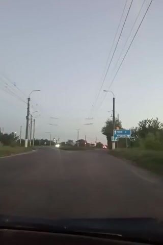 The video shows a view from inside a car driving on an asphalt road. The sky is a light gray, suggesting dawn or dusk. Power lines stretch across the upper portion of the frame. On the right side of the road, there is a sign pointing towards "Rivne" to the left and "Luts’k" straight ahead. Streetlights line the sides of the road. In the distance, traffic lights are visible, showing red. The car is moving forward, towards the traffic lights. Some greenery and trees border the side of the road. The car’s dashboard is visible at the bottom of the frame. There is music playing in the background.