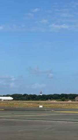An airplane with a white fuselage and a red and white checkered tail, marked "VOLOTEA," taxis from right to left across an airport runway with yellow markings. An all-white airplane is partially visible and stationary on the far left. In the background, a line of green trees and a distant tower are visible under a blue sky with scattered white clouds.