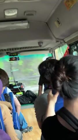 The video shows the interior of a minibus with passengers, likely on a tour or transport to an activity. Passengers are wearing blue life vests, suggesting they may be going on a water-based excursion. The minibus is moving, as evidenced by the blurred scenery visible through the windshield. The vehicle's dashboard and interior are visible, along with the heads and backs of several passengers. The lighting inside is bright and possibly overcast outside. The time displayed on the rearview mirror indicates it is 11:23.
