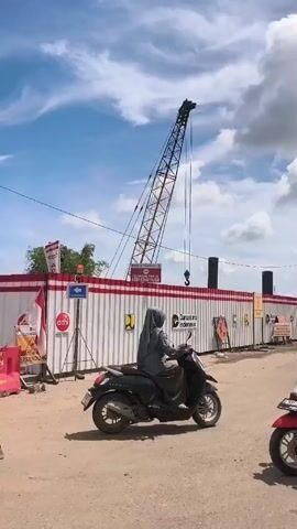 The video shows a construction site on a partly cloudy day. In the background, there's a tall crane. A long white fence with a red stripe runs across the scene. The fence has logos, including "Adhi" and "Danantara Indonesia," and signs posted on it. People on motorbikes are seen driving past the construction site. The road is unpaved and appears to be dusty.