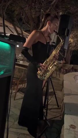 A woman in a black dress and gloves plays a saxophone in an outdoor setting at night. She is standing next to a speaker and a stand for the instrument. Trees and chairs are visible in the background.