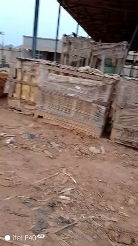 The camera pans across a storage area with a dirt ground.  Wooden pallets wrapped in clear plastic and wooden crates are stacked. Red, white, and blue plastic barrels are stacked in the background. Metal rods and coils are scattered near the center.  A corrugated metal roof covers the area.  Hills covered in vegetation are visible in the distance.