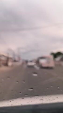 The video captures a daytime road scene from the perspective of a vehicle driving on a paved road in Grong-Grong, Indonesia. The view is through a windshield with noticeable water droplets or smudges.

The sky is overcast and heavily clouded, casting a diffused light over the scene. Two prominent white vehicles are driving ahead of the camera vehicle. In the lead is a white hatchback car, appearing to be a Honda Jazz/Fit model, bearing the license plate "BL 1464 AAH". Following it closely is a white ambulance van. The ambulance is clearly marked at the rear with "EMERGENCY" in red capital letters at the top and "AMBULANCE" in red capital letters at the bottom, along with a red cross symbol. Smaller, blurry text below "EMERGENCY" appears to indicate "RUMAH SAKIT UMUM DAERAH" (Regional General Hospital) and its license plate reads "BL 9298 AE".

The road has at least two lanes in the direction of travel, separated by a broken white line. Along the left side of the road, several low-rise buildings are visible, some appearing commercial or residential, alongside numerous utility poles and overhead power lines. Sparse vegetation can be seen on the roadside. All vehicles are in motion, typical of traffic on a provincial road.