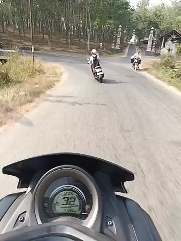 The video shows a first-person perspective of someone riding a motorcycle on a paved road. The rider's dashboard is visible, displaying a speed of 23 km/h. The road is bordered by trees and dry grass. In the distance, other motorcycles and a black van are seen. One motorcycle in the distance has a person wearing a helmet and a backpack, while another motorcycle has two people on it. The black van appears to be parked on the side of the road. The environment suggests a rural or semi-rural setting with a clear sky.