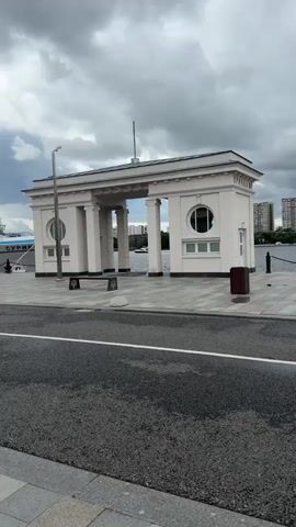 The video opens with a stationary shot of a light-colored building with arched entrances and circular windows, resembling a gateway or a pavilion. The building is situated on a paved area next to a body of water. A white boat with the inscription "ВАСИЛИЙ СУРИКОВ" is docked to the left of the building. The camera then pans left, revealing a large white passenger ship docked along the waterfront. The ship has multiple decks and numerous windows. As the camera continues to pan, it shows a curved roadway in the foreground with a white line marking its center. The background reveals a cloudy sky and buildings in the distance. The overall scene depicts a waterfront area with a passenger ship and architectural elements.